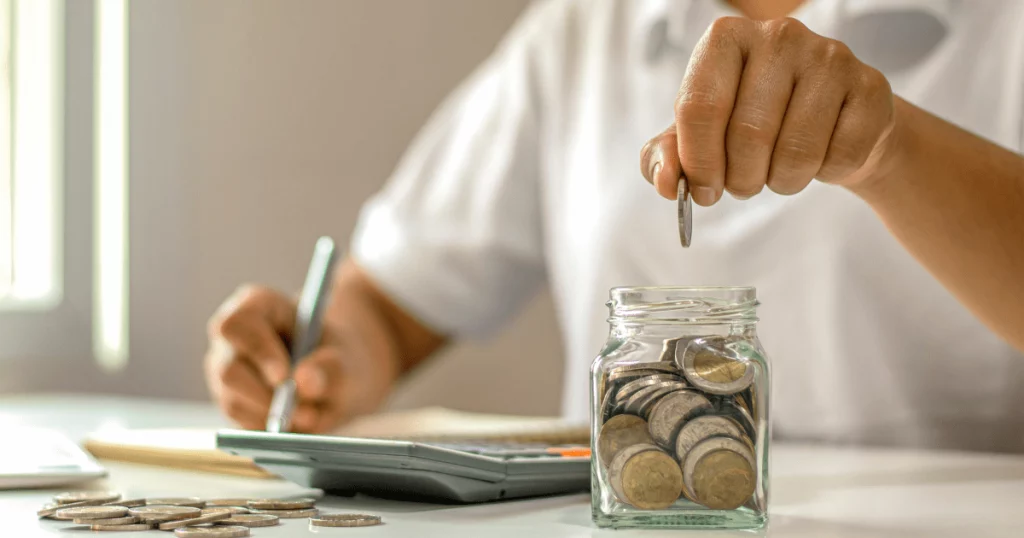 Mão depositando moedas em um pote transparente, representando economia e planejamento para aposentar sem contribuição.
