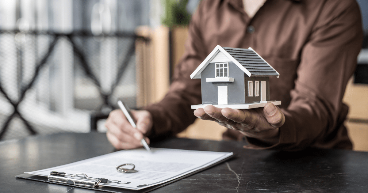 Mão segurando uma miniatura de casa enquanto assina documentos, representando o processo de usucapião para regularização de imóveis.